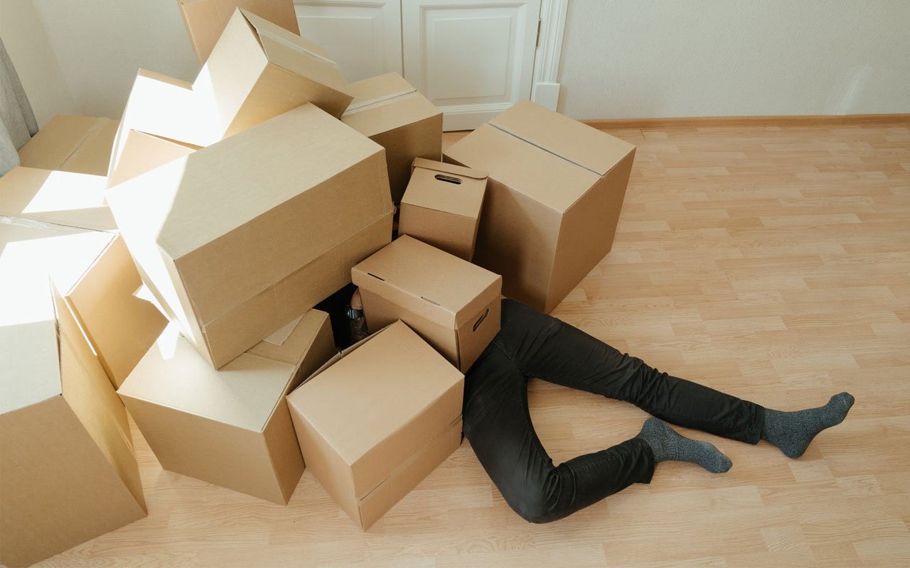 A person is lying on the floor, partially buried under a pile of cardboard boxes in a bright room.