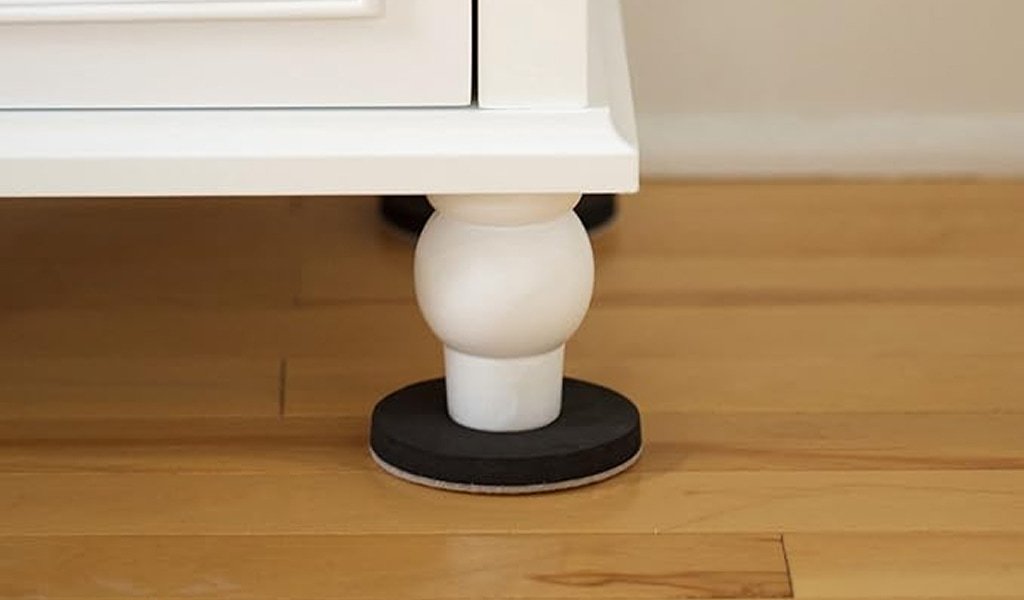 White furniture leg resting on a black round pad, positioned over light wooden flooring. The design is simple and functional.