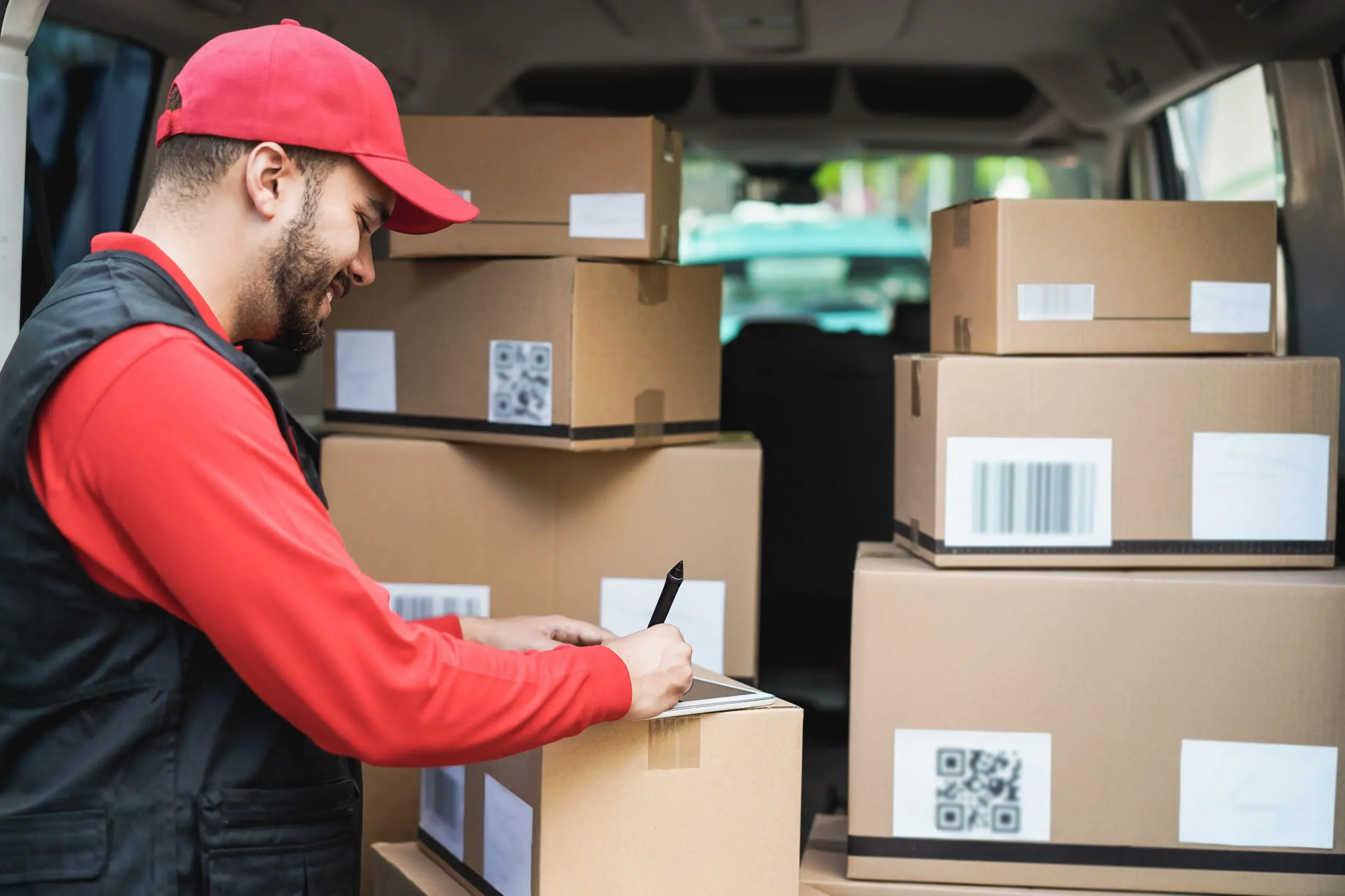 A delivery worker in a red shirt and cap is marking a box among several brown cardboard packages in a vehicle's cargo area.
