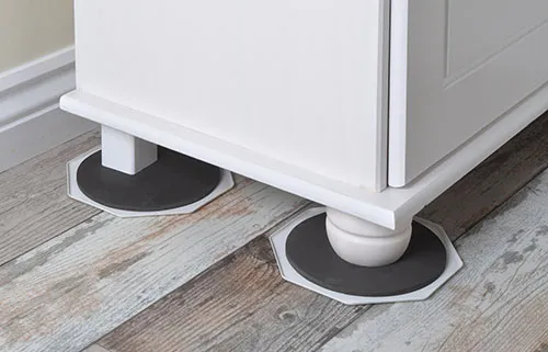 A white cabinet with rounded feet resting on octagonal black pads, positioned on wooden flooring.