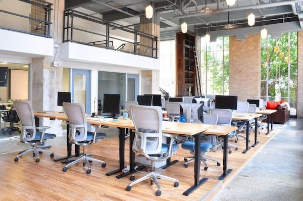 Modern open office space featuring rows of desks with ergonomic chairs, computers, and large windows allowing natural light.