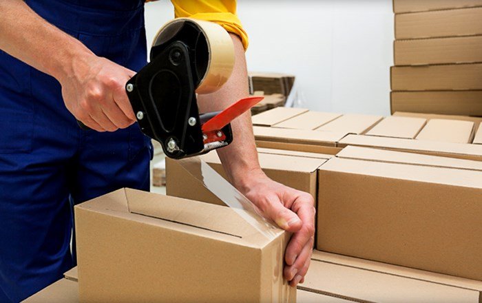 A worker using a tape dispenser to seal a cardboard box, surrounded by stacked boxes in a warehouse setting.