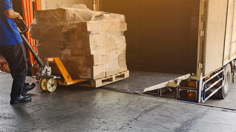 A worker uses a pallet jack to unload a wrapped pallet of boxes from a truck's loading ramp in a warehouse setting.