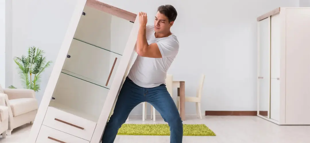 A person is struggling to lift a large, white cabinet in a bright, modern room with minimal furniture and greenery.