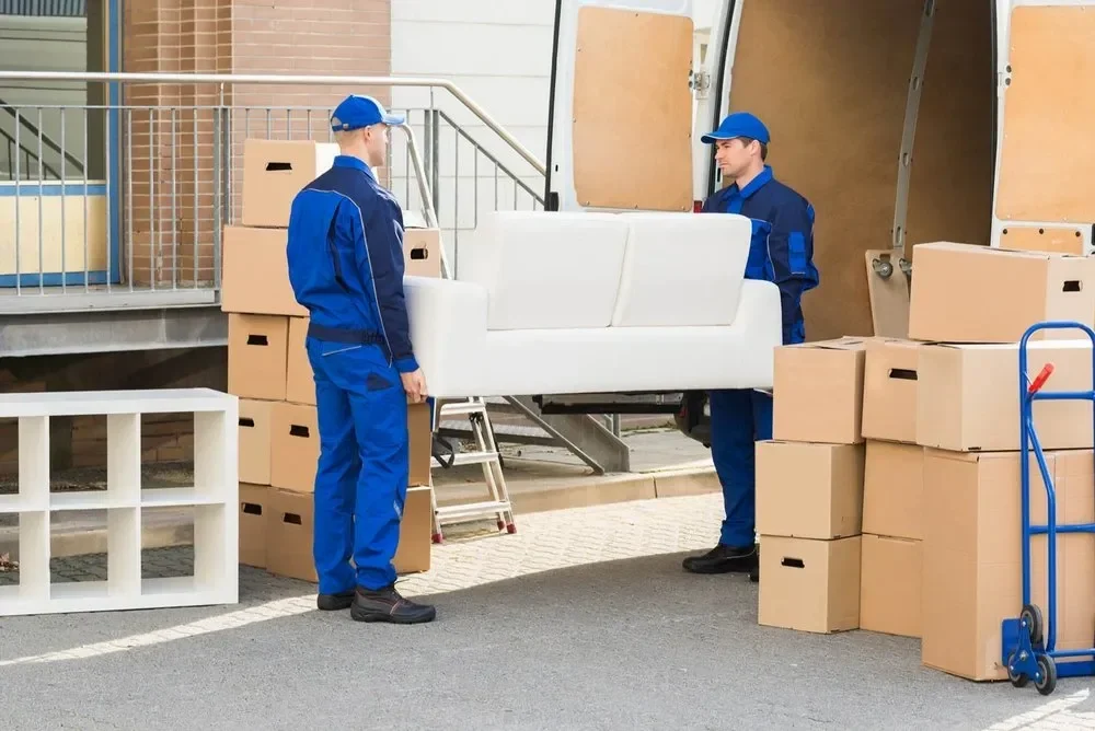 Two movers in blue uniforms carry a white sofa, surrounded by moving boxes and a dolly, in front of a van.