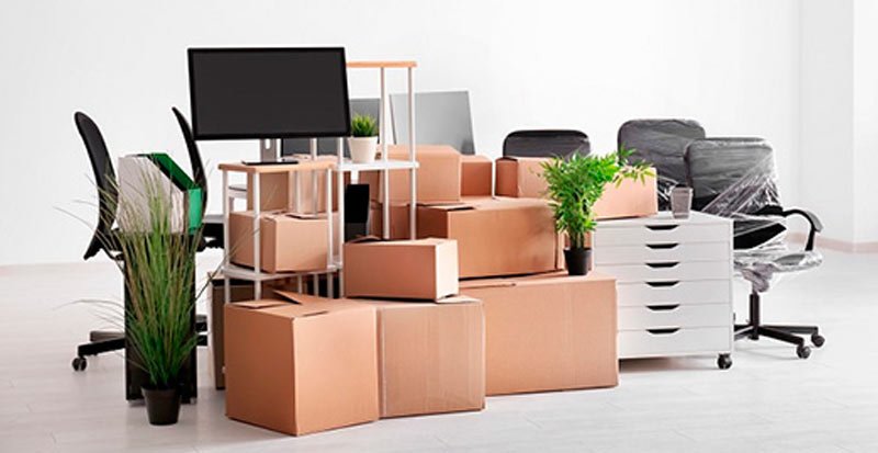 An office space filled with stacked cardboard boxes, a monitor on a stand, and potted plants, surrounded by office chairs.